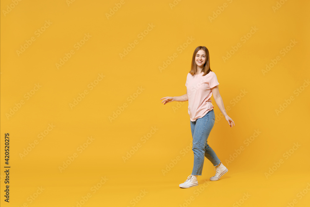 Full length of young friendly smiling student woman 20s with nude make up wearing casual basic pastel pink t-shirt, jeans looking camera, walking going isolated on yellow background studio portrait. Full length of young friendly smiling student woman 20s with nude make up wearing casual basic pastel pink t-shirt, jeans looking camera, walking going isolated on yellow background studio portrait.