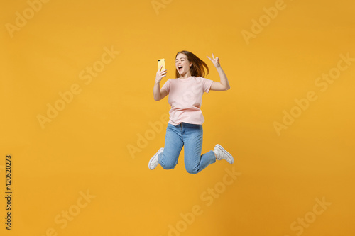 Full length of young expressive friendly overjoyed woman 20s in basic pastel pink t-shirt jump high holding mobile cell phone show victory v-sign gesture isolated on yellow background studio portrait. © ViDi Studio
