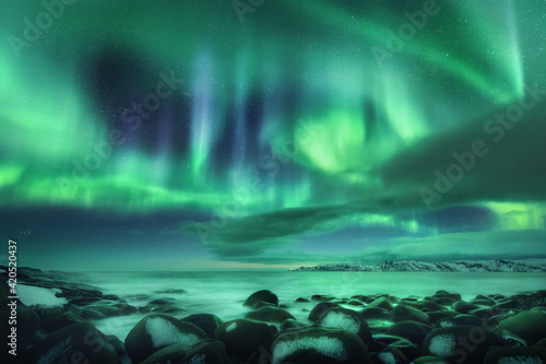 Aurora borealis over ocean. Northern lights in Teriberka, Russia. Starry sky with polar lights and clouds. Night winter landscape with bright aurora, stars, sea, snowy stones in blurred water. Travel © den-belitsky