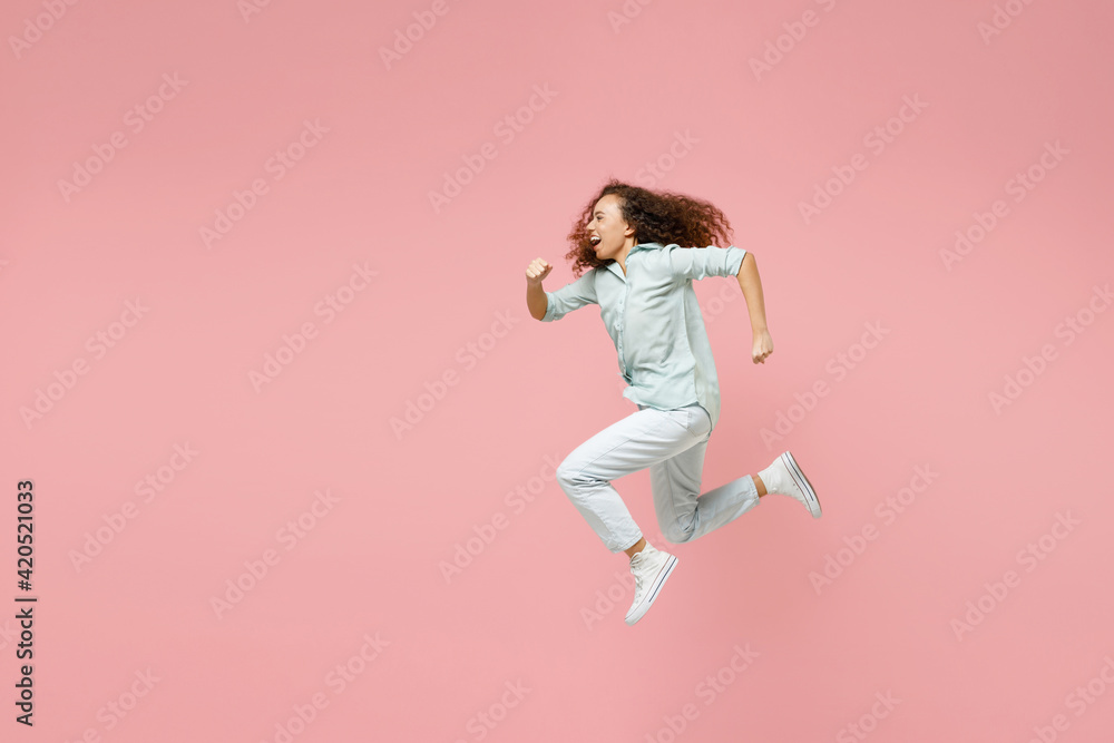 Full length side view young black african fun happy smiling energetic sporty excited woman 20s in blue shirt jump high running fast hurrying up isolated on pastel pink color background studio portrait Full length side view young black african fun happy smiling energetic sporty excited woman 20s in blue shirt jump high running fast hurrying up isolated on pastel pink color background studio portrait
