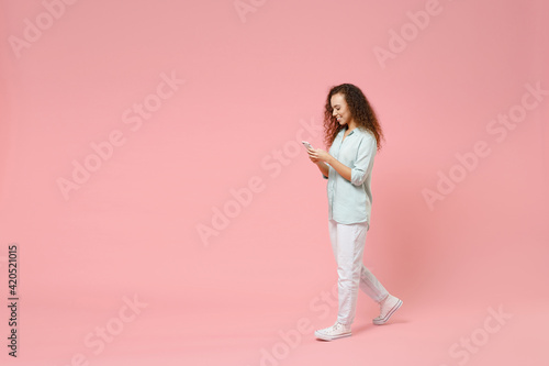 Full length young black african fun happy smiling curly student woman 20s in blue shirt holding mobile cell phone chatting browsing internet walk isolated on pastel pink background studio portrait.