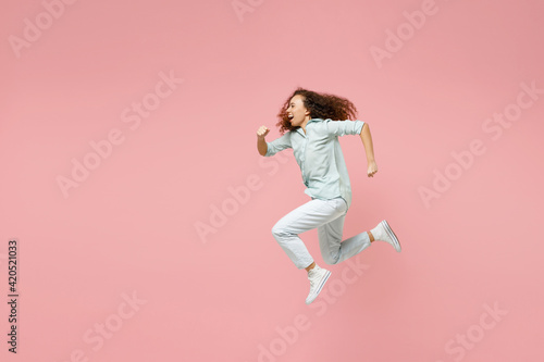 Full length side view young black african fun happy smiling energetic sporty excited woman 20s in blue shirt jump high running fast hurrying up isolated on pastel pink color background studio portrait