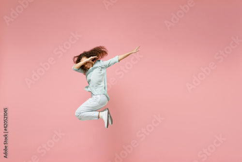 Full length young black african fun woman 20s wear blue shirt doing dab hip hop dance hands move gesture youth sign hiding covering face jump high isolated on pastel pink background studio portrait
