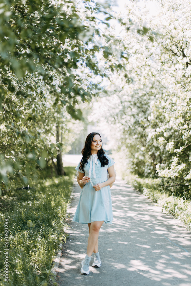 Gentle beautiful young happy brunette woman in a blue dress with a straw hat smiling relaxing enjoying a walk in the park in spring blooming apple trees outdoors, selective focus Gentle beautiful young happy brunette woman in a blue dress with a straw hat smiling relaxing enjoying a walk in the park in spring blooming apple trees outdoors, selective focus