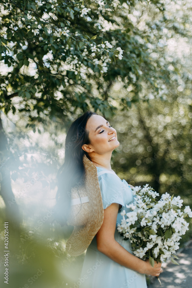 Gentle beautiful young happy brunette woman in a blue dress with a straw hat smiling relaxing enjoying a walk in the park in spring blooming apple trees outdoors, selective focus Gentle beautiful young happy brunette woman in a blue dress with a straw hat smiling relaxing enjoying a walk in the park in spring blooming apple trees outdoors, selective focus