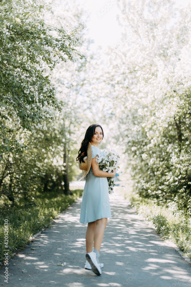 Gentle beautiful young happy brunette woman in a blue dress with a straw hat smiling relaxing enjoying a walk in the park in spring blooming apple trees outdoors, selective focus Gentle beautiful young happy brunette woman in a blue dress with a straw hat smiling relaxing enjoying a walk in the park in spring blooming apple trees outdoors, selective focus
