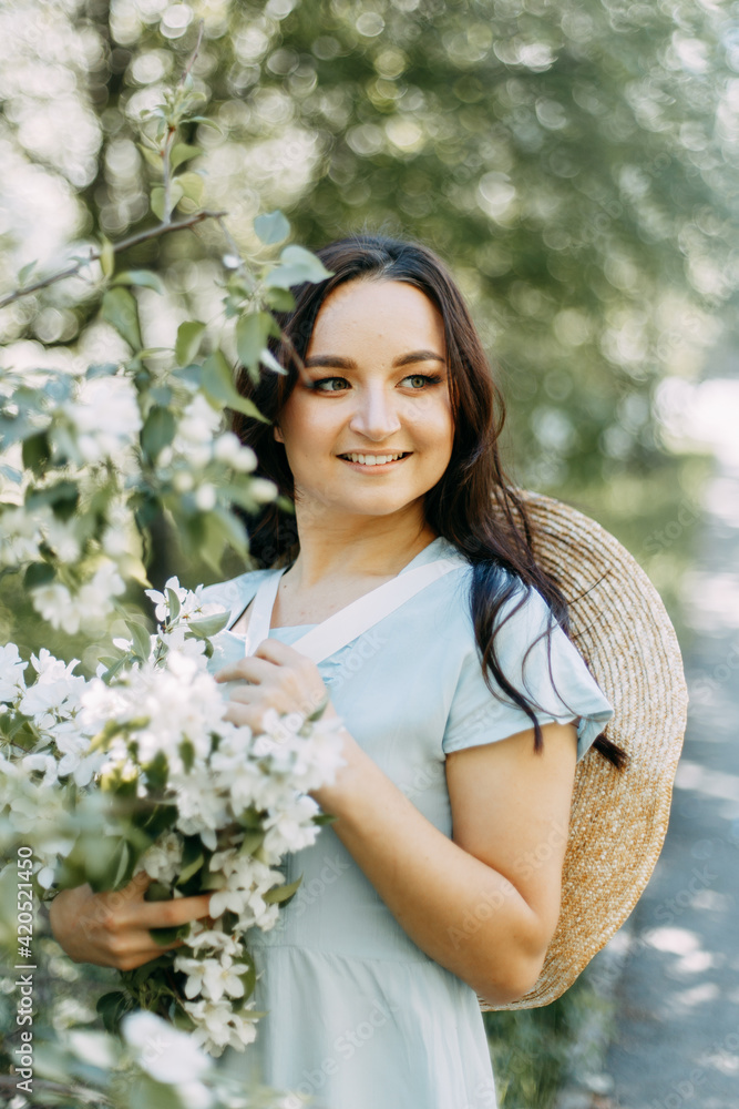 Gentle beautiful young happy brunette woman in a blue dress with a straw hat smiling relaxing enjoying a walk in the park in spring blooming apple trees outdoors, selective focus Gentle beautiful young happy brunette woman in a blue dress with a straw hat smiling relaxing enjoying a walk in the park in spring blooming apple trees outdoors, selective focus
