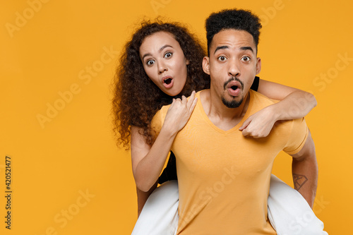 Young couple two friends together family african shocked surprised man woman with open mouth in black t-shirt giving piggyback ride to joyful sit on back isolated on yellow background studio portrait.
