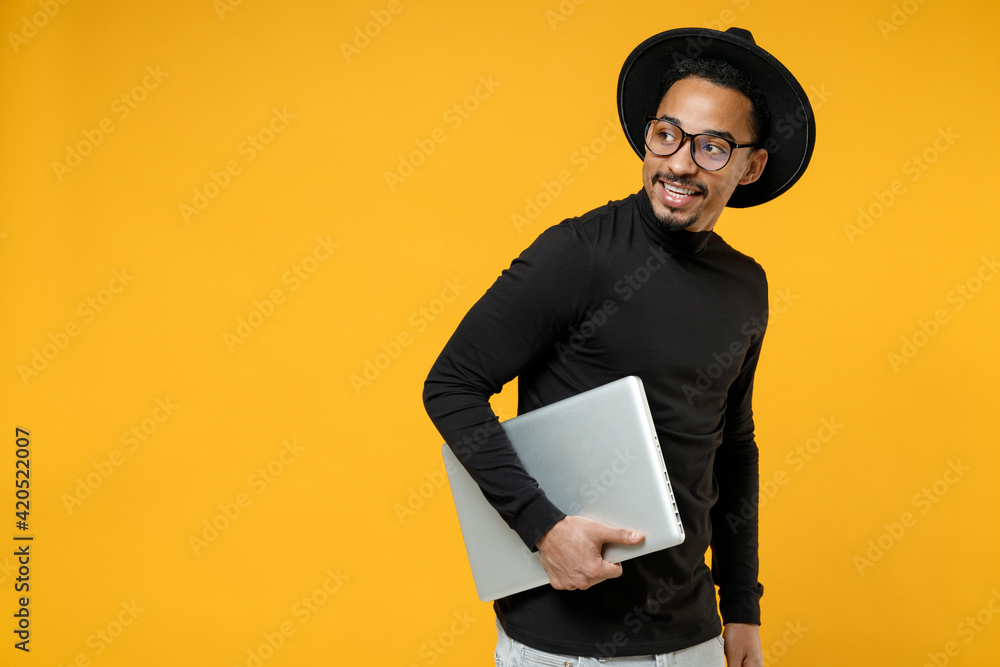 Side view of young freelancer smiling copywriter african man 20s wear stylish black shirt hat eyeglasses walk with closed laptop pc computer look aside isolated on yellow background studio portrait