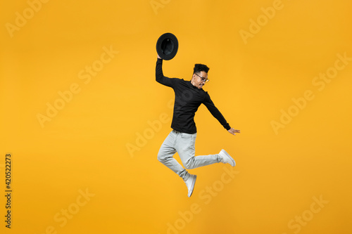 Full length of young active fashionable overjoyed fun happy african man in stylish black shirt eyeglasses jump high with outstretched hands took off hat isolated on yellow background studio portrait.