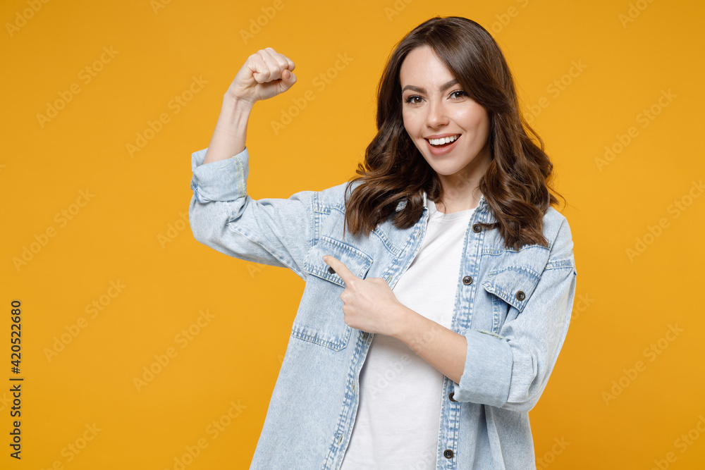 Young brunette fun strong sporty fitness woman 20s in denim shirt white t-shirt point index finger on biceps muscles on hand demonstrating strength power isolated on yellow background studio portrait. Young brunette fun strong sporty fitness woman 20s in denim shirt white t-shirt point index finger on biceps muscles on hand demonstrating strength power isolated on yellow background studio portrait.