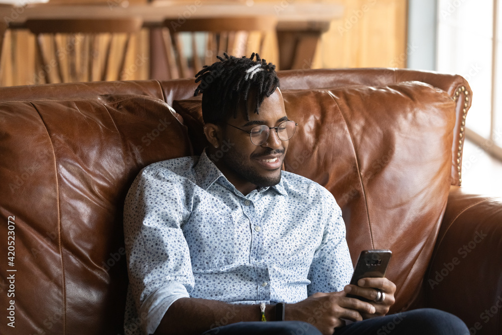 Fototapeta premium Passionate gamer. Joyful millennial african guy student relax at home sit on couch play smartphone based online game. Young black man hipster addicted to internet spend free time using mobile phone