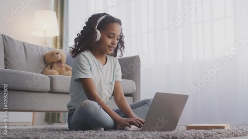 Teen black girl in headphones typing on laptop, communicating with teacher via video chat, sitting on floor at home photo