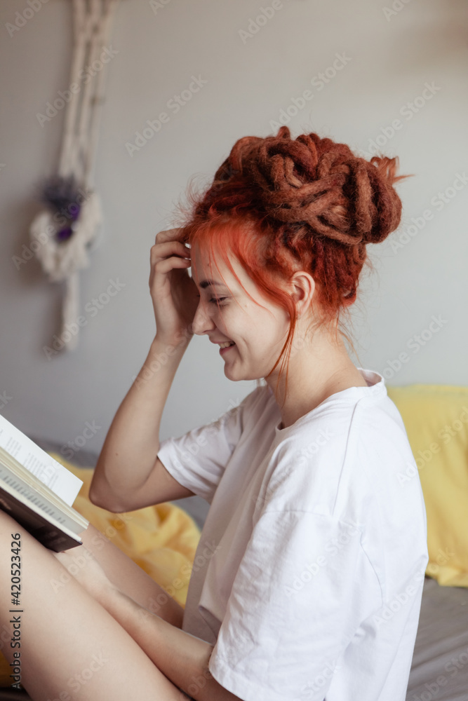 Very pretty redhead girl with dreads on bed in bedroom in the morning. Young woman reading a book sitting in bed. A feminine, gentle girl. Very pretty redhead girl with dreads on bed in bedroom in the morning. Young woman reading a book sitting in bed. A feminine, gentle girl.
