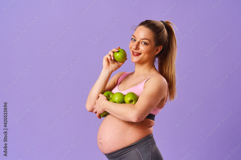 attractive, happy pregnant woman holds fresh organic green apples while standing on purple background. expectant mother eats an apple. Healthy, proper nutrition. Vegetarian, dietary food. Vitamins. attractive, happy pregnant woman holds fresh organic green apples while standing on purple background. expectant mother eats an apple. Healthy, proper nutrition. Vegetarian, dietary food. Vitamins.