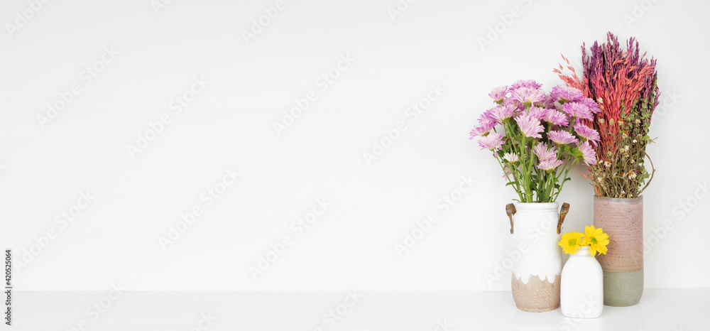 Fototapeta premium Home decor on a shelf. Colorful flowers and branches in vases. White shelf against a white wall. Banner with copy space.