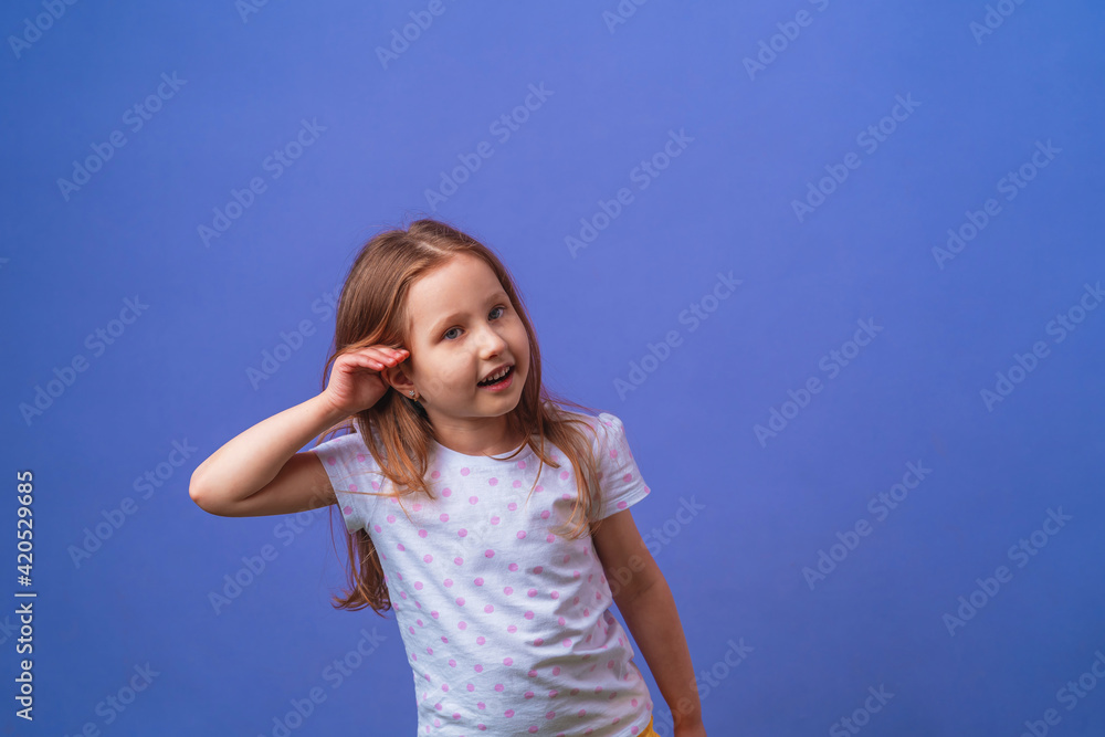 charming little girl smiles with her hand over her ear while listening and hearing rumors or gossip on a purple isolated background. Child eavesdropping