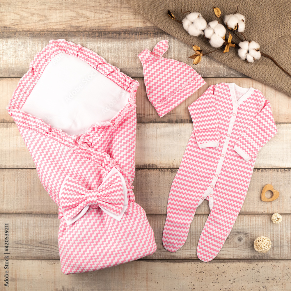 Clothing for a newborn at discharge from the maternity hospital. A blanket, a bow, a cap and a jumpsuit for a newborn baby of pink and white color made of natural cotton, on wooden boards. Top view.