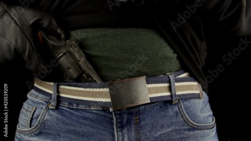 Close-up, man with leather gloves pulls out a gun from the denim belt. Man hiding gun under pants, isolated on black background.