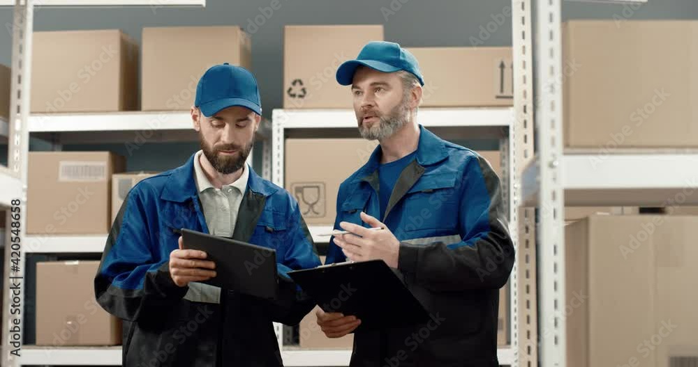 Two couriers in uniform stand in warehouse with cardboard boxes and holding folders with documents distribute parcels for delivery. Two couriers in uniform stand in warehouse with cardboard boxes and holding folders with documents distribute parcels for delivery.