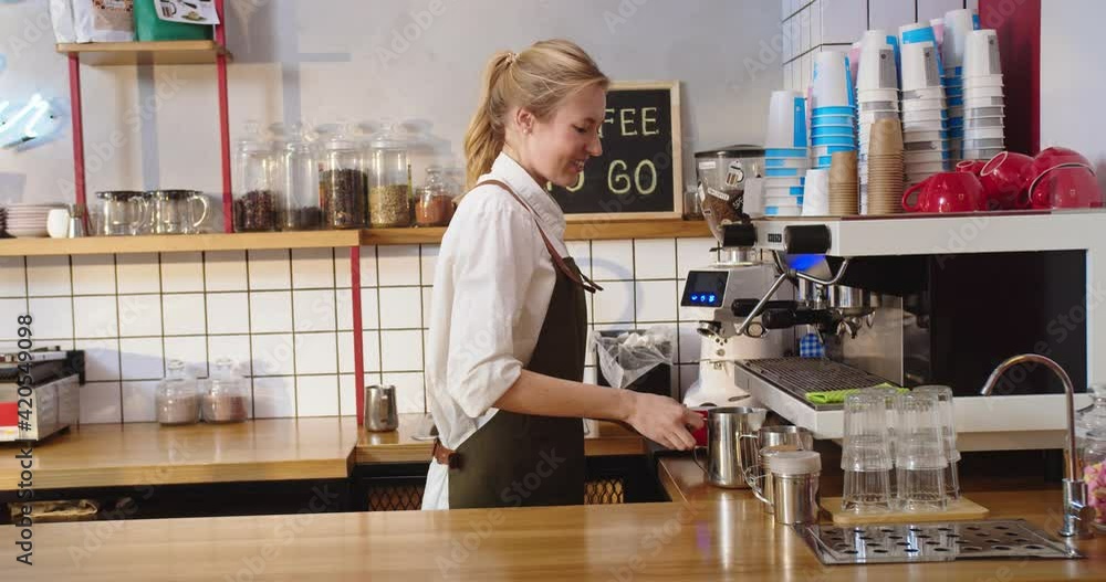 Beautiful Caucasian woman in apron making beverages on coffee machine. Young female barista lady holding cup, looking at camera and smiling. Career, business, part-time job concept. Beautiful Caucasian woman in apron making beverages on coffee machine. Young female barista lady holding cup, looking at camera and smiling. Career, business, part-time job concept.