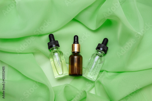 Three cosmetic glass bottles with oil, serum and hyaluronic acid with pipette on green silk fabric with pleats close up, flat lay, face and body care concept, women's anti-aging cosmetics, copy space photo