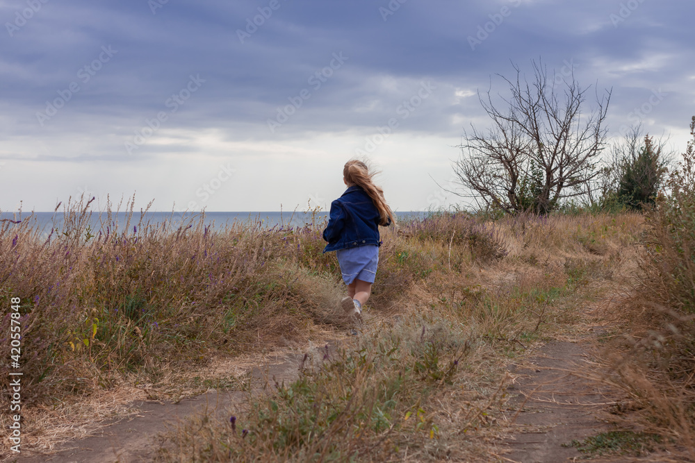 Adorable little girl in denim jacket back view on a hill with sea landscape view. Stylish child with long blonde hair on countryside cloudy sky background. Outdoor walking rural road trip faceless. Adorable little girl in denim jacket back view on a hill with sea landscape view. Stylish child with long blonde hair on countryside cloudy sky background. Outdoor walking rural road trip faceless.