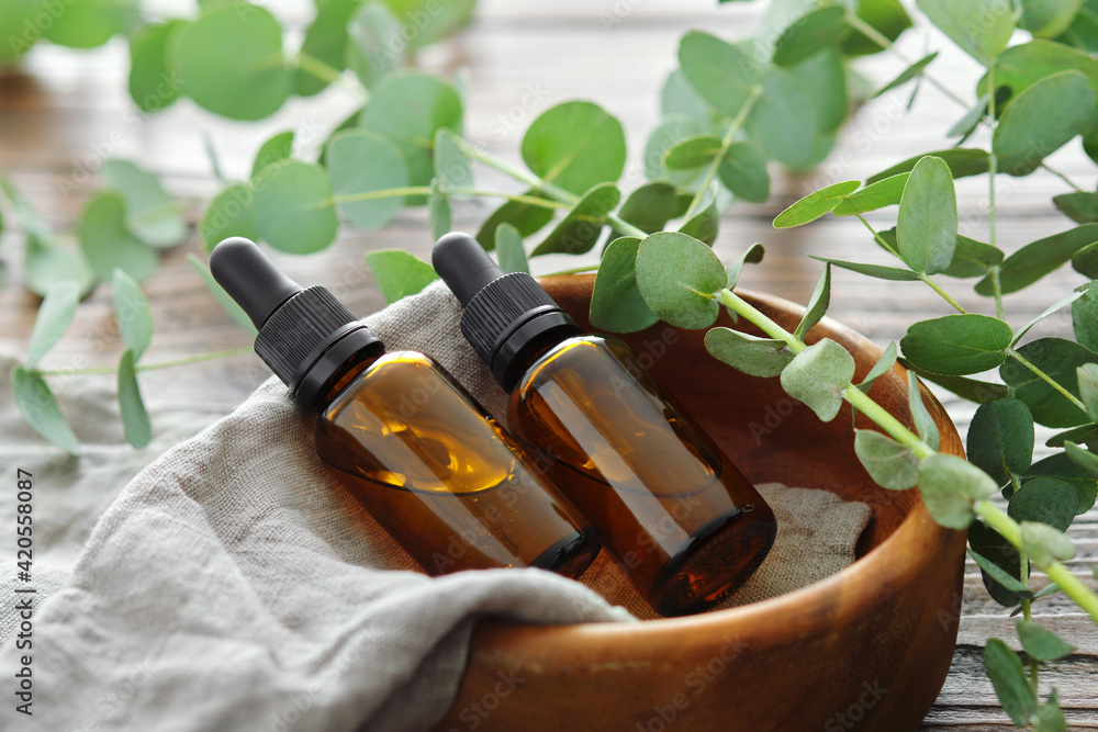 Two bottles of eucalyptus essential oil in wooden bowl and eucalyptus twigs on table. - obrazy, fototapety, plakaty 