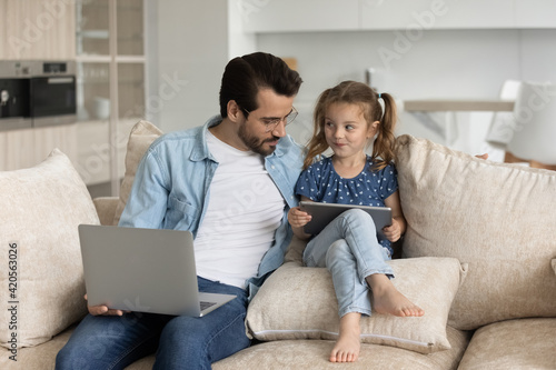 Cute little Caucasian girl child and young father relax on sofa at home use electronic devices laptop and tablet. Dad and small 7s daughter rest together on couch with gadgets. Technology concept.