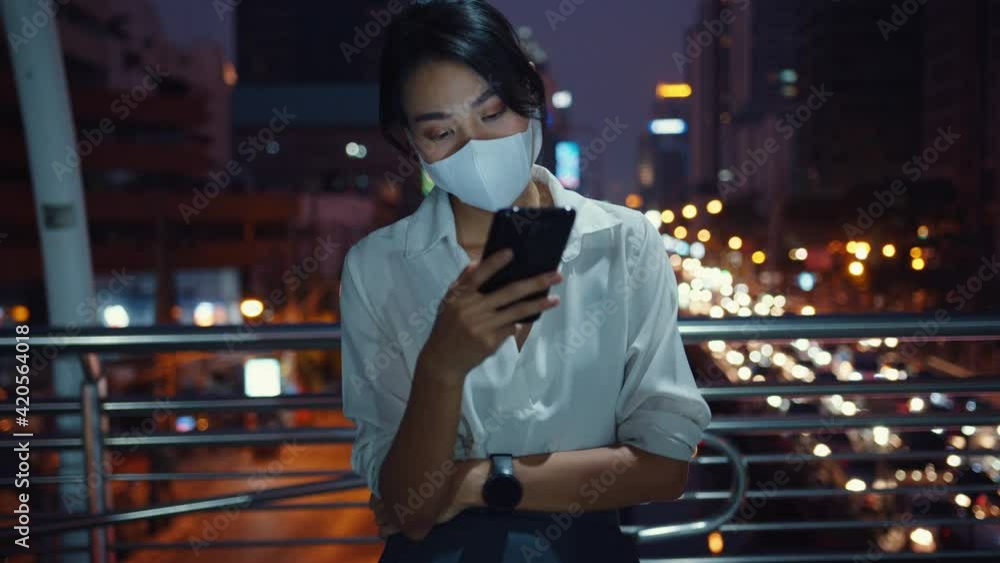 Young Asia businesswoman in fashion clothes wearing face mask using smart phone typing text message while stand outdoor in urban city at night. Social distancing to prevent spread of COVID-19 concept. Young Asia businesswoman in fashion clothes wearing face mask using smart phone typing text message while stand outdoor in urban city at night. Social distancing to prevent spread of COVID-19 concept.
