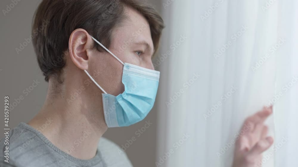 Close-up face of depressed isolated Caucasian man in coronavirus face mask looking out the window thinking. Side view portrait of frustrated brunette ill guy on Covid-19 lockdown. Stress on pandemic. Close-up face of depressed isolated Caucasian man in coronavirus face mask looking out the window thinking. Side view portrait of frustrated brunette ill guy on Covid-19 lockdown. Stress on pandemic.