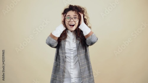 Shocked, excited young woman with her hands in her hair says wow looking curiously at the camera Cretaceous in gray jacket and white shirt, with glasses posing isolated on a beige background in the