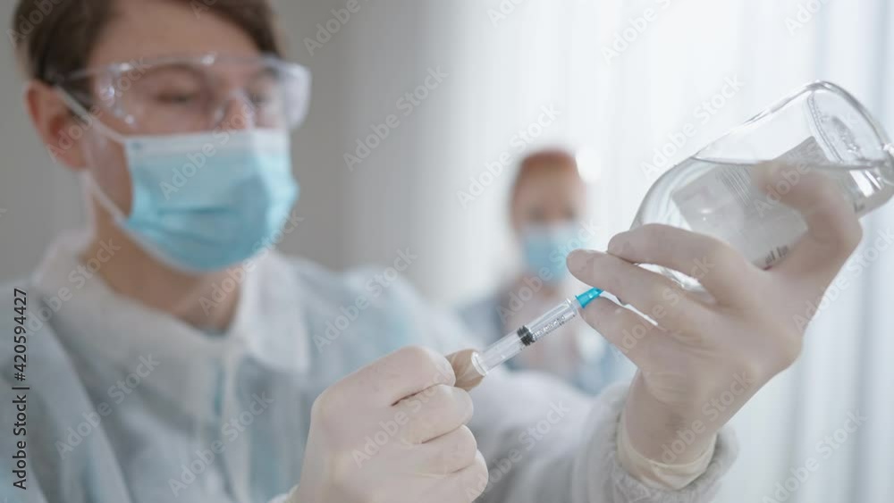 Male doctor filling syringe with liquid medication as blurred nurse standing at background. Professional Caucasian man and woman preparing Covid-19 vaccination in hospital indoors. Male doctor filling syringe with liquid medication as blurred nurse standing at background. Professional Caucasian man and woman preparing Covid-19 vaccination in hospital indoors.