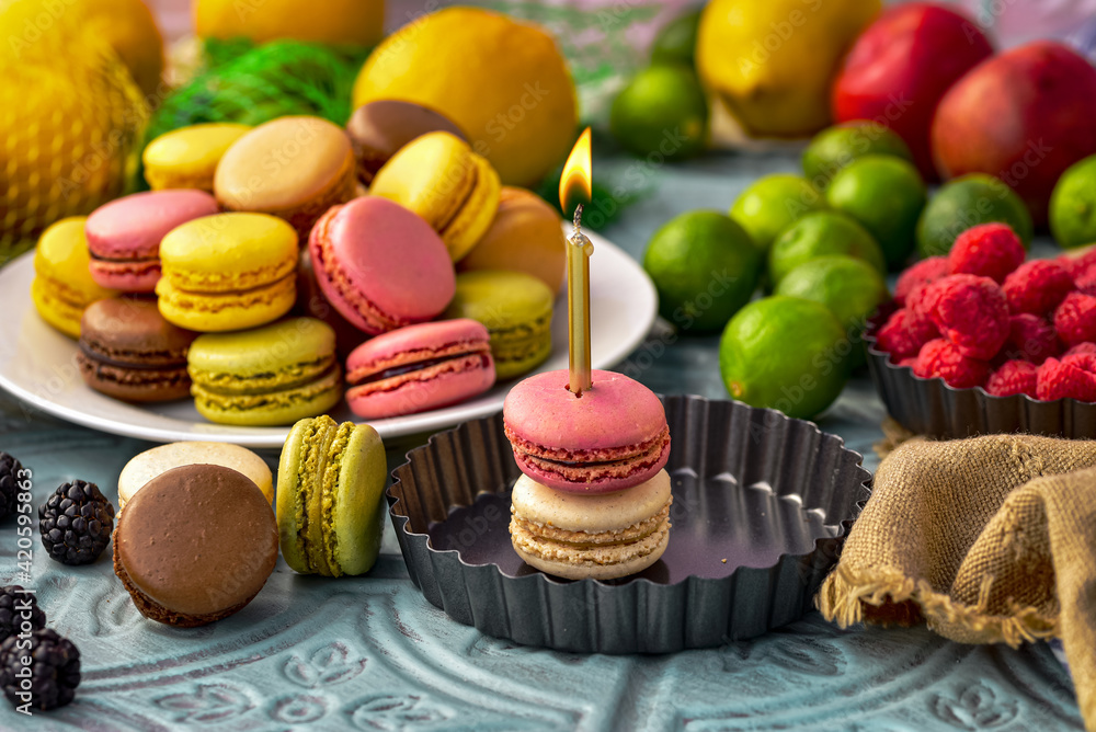 tabletop still life with fruit, plate of assorted fruit flavor macaron cookies, and two macarons stacked with gold birthday candle lit with flame tabletop still life with fruit, plate of assorted fruit flavor macaron cookies, and two macarons stacked with gold birthday candle lit with flame