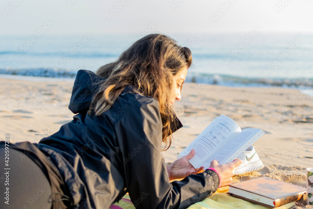 Joven mujer leyendo un libro al amanecer junto al mar.