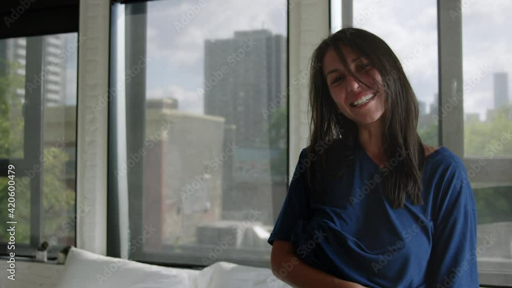 Young adult woman looks towards camera and smiles in sunny bedroom on summer morning - wide shot Young adult woman looks towards camera and smiles in sunny bedroom on summer morning - wide shot