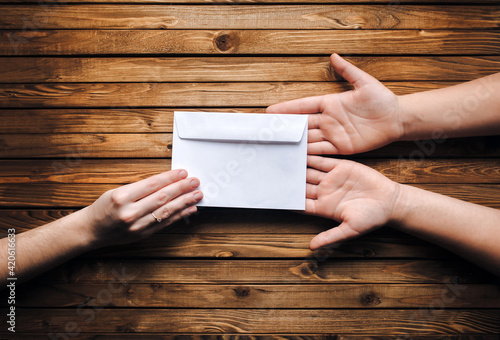 White small paper envelope (letter) in the hands of two people on brown wooden background. The concept of writing, online donation, email. The Internet.