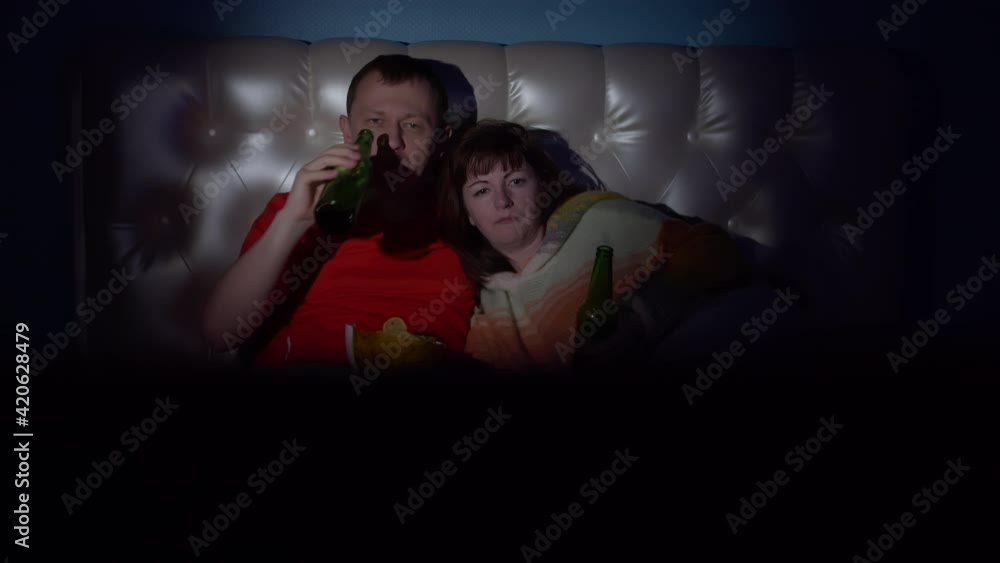 happy man and woman lie on the bed drink beer with chips and watch tv, smiling