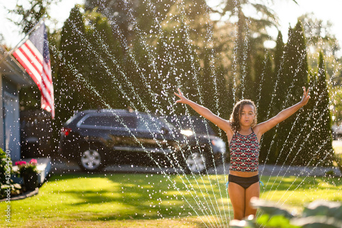 Curly haired girl stands in sprinkler with arms raised photo