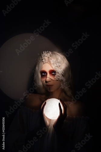 concept portrait of a beautiful young woman on a black background with bright dark makeup and the moon in her hands, who plays the role of a priestess, spirit or ghost on Halloween