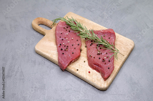 Raw beef steak with spices and rosemary on gray background. Fresh meat rib eye steak. Cooking beef steak fillets. Slices of medium rare roast beef meat on wooden cutting board. Top view.