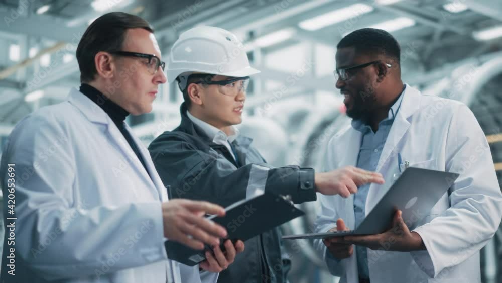 Team of Diverse Professional Heavy Industry Engineers Wearing Safety Uniform and Hard Hats Working on Laptop Computer. Technician and Workers Talking on a Meeting in a Factory Facility. Team of Diverse Professional Heavy Industry Engineers Wearing Safety Uniform and Hard Hats Working on Laptop Computer. Technician and Workers Talking on a Meeting in a Factory Facility.