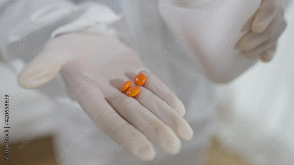 Close-up of female hands in white gloves pouring orange pills from container. Unrecognizable nurse or doctor showing medication in palm. Medicine and treatment concept. Close-up of female hands in white gloves pouring orange pills from container. Unrecognizable nurse or doctor showing medication in palm. Medicine and treatment concept.