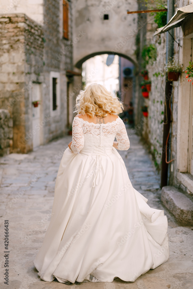 The bride walks along a beautiful narrow street of the old town of Perast with brick houses, back view 