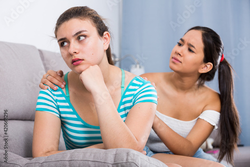 Teen girl trying to calm her female friend sitting on sofa and apologize after dispute