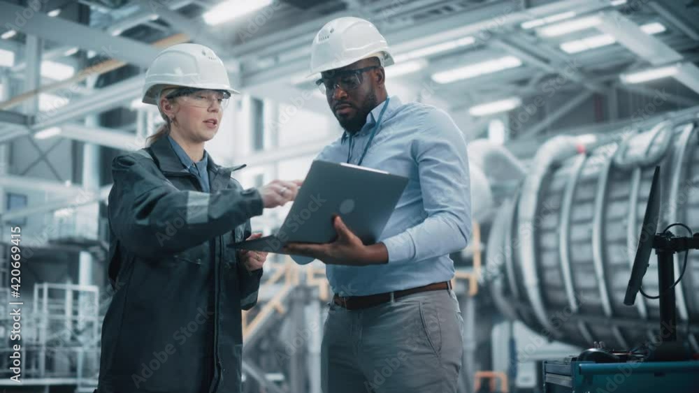 Two Diverse Professional Heavy Industry Engineers Wearing Safety Uniform and Hard Hats Working on Laptop Computer. African American Technician and Female Worker Talking on a Meeting in a Factory. Two Diverse Professional Heavy Industry Engineers Wearing Safety Uniform and Hard Hats Working on Laptop Computer. African American Technician and Female Worker Talking on a Meeting in a Factory.