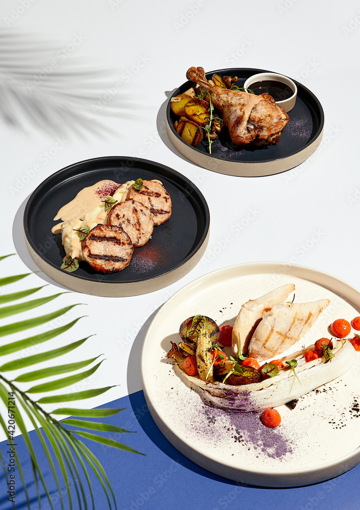 Various meat poultry dishes on white and blue table. Chicken and turkey poultry food plate. Restaurant plate in modern food style. Sunlight with harsh shadow of palm leaves. Various meat poultry dishes on white and blue table. Chicken and turkey poultry food plate. Restaurant plate in modern food style. Sunlight with harsh shadow of palm leaves.