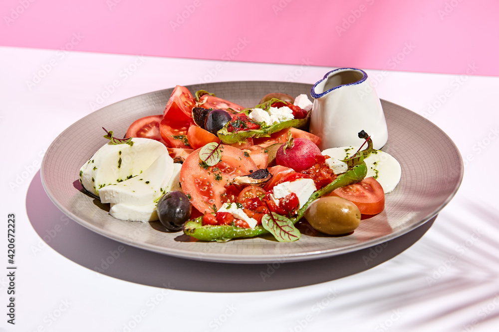 Tomato, olives and mozzarella salad . Restaurant appetizer plate on white table with pink wall. Day sunlight with hard shadow of fern palm leaves. Summer or spring restaurant food concept