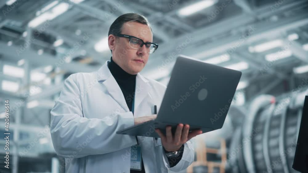Professional Male Heavy Industry Engineer/Worker Working on a Laptop Computer. Confident Caucasian Industrial Specialist Wearing White Laboratory Robe and Safety Glasses in a Factory Facility. Professional Male Heavy Industry Engineer/Worker Working on a Laptop Computer. Confident Caucasian Industrial Specialist Wearing White Laboratory Robe and Safety Glasses in a Factory Facility.