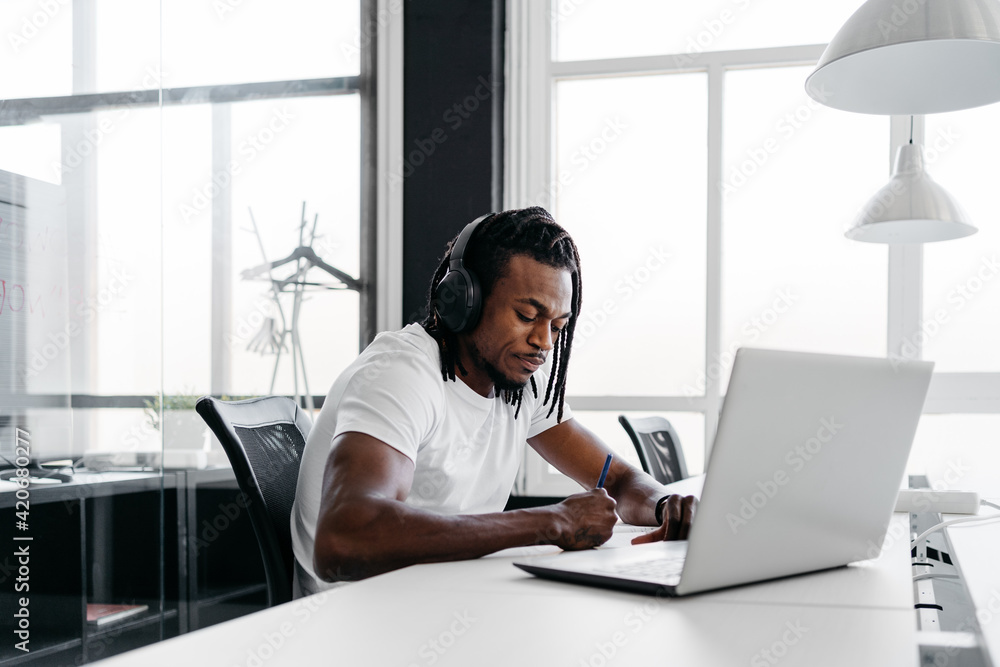 Black man wearing headphones writing on a notebook while working with his laptop in a bright modern coworking Black man wearing headphones writing on a notebook while working with his laptop in a bright modern coworking
